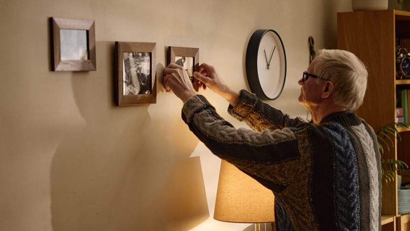 An older man wearing glasses hangs a framed photo on a wall next to other pictures—a perfect example of how to make a senior apartment feel like home. A round clock, lamp, and bookshelf are also visible in the cozy room.