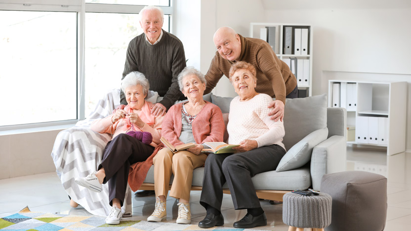Five smiling elderly people, three women seated on a couch and two men standing behind them, pose together in a bright living room with bookshelves and large windows—showing how seniors make new friends by enjoying social gatherings and shared spaces.