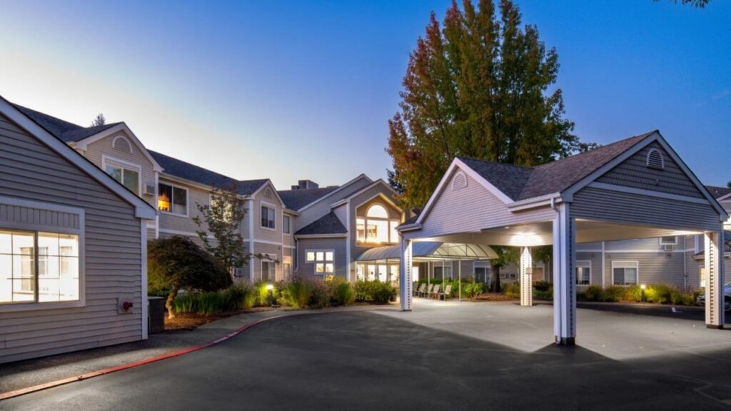 Exterior view of a large, well-lit building with a covered driveway, large windows, and landscaping, photographed at dusk with a tree in the background and a clear blue sky.