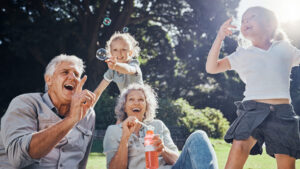 Two older adults and two children play with bubbles outside on a sunny day. The group is smiling and laughing, surrounded by greenery, enjoying each other’s company and the cheerful atmosphere—capturing one of many family activity ideas in senior living communities.