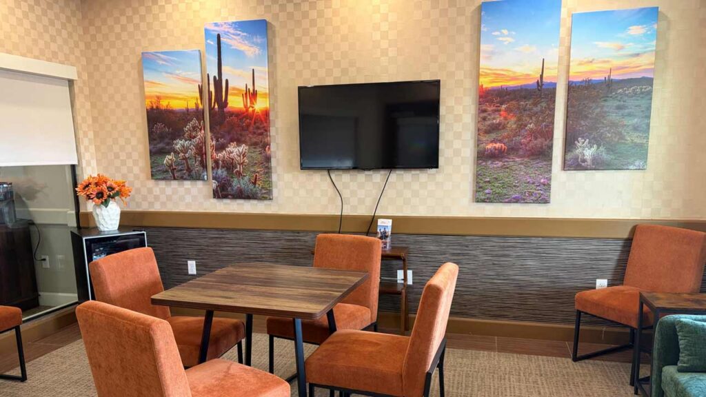 A small room with orange chairs around a wooden table, desert landscape art on the walls, a flat-screen TV, and a vase of flowers on a counter.