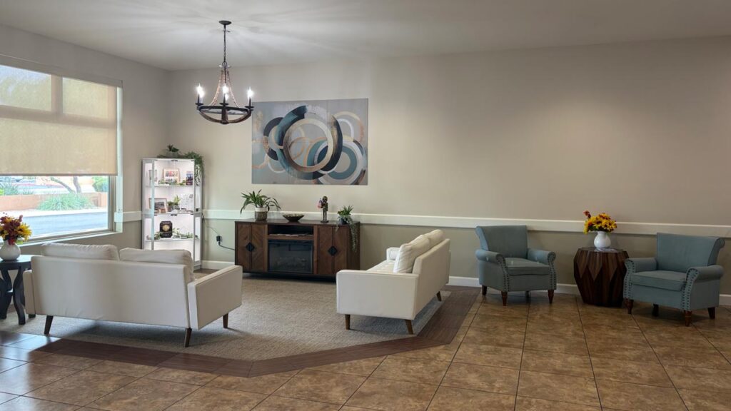 A bright, modern lounge with white sofas, blue armchairs, sunflowers, a chandelier, tiled floor, wall art, and large window. Shelves and plants decorate the space, with light streaming in.