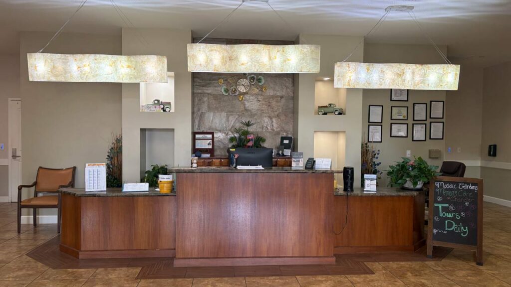Reception desk in a lobby with three rectangular hanging lights, decorative plants, a wall display with awards and toy cars, a blackboard with tour information, and a chair in the corner.