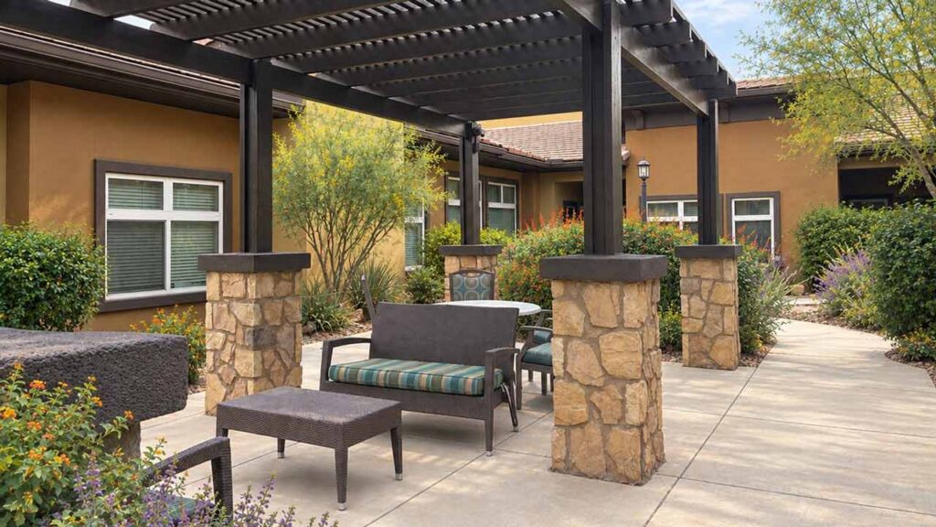 Outdoor patio area with cushioned chairs and a bench under a wooden pergola, surrounded by stone pillars, plants, and walkway near a tan building with white-trimmed windows.