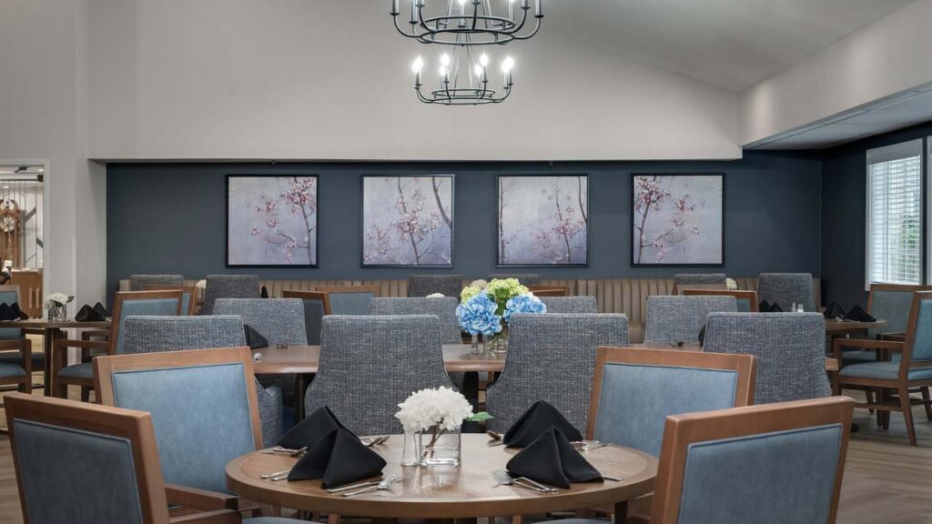 A modern dining room with gray chairs, round wooden tables set with black napkins, and flower centerpieces. Three framed cherry blossom prints hang on a dark accent wall. A chandelier hangs from the ceiling.