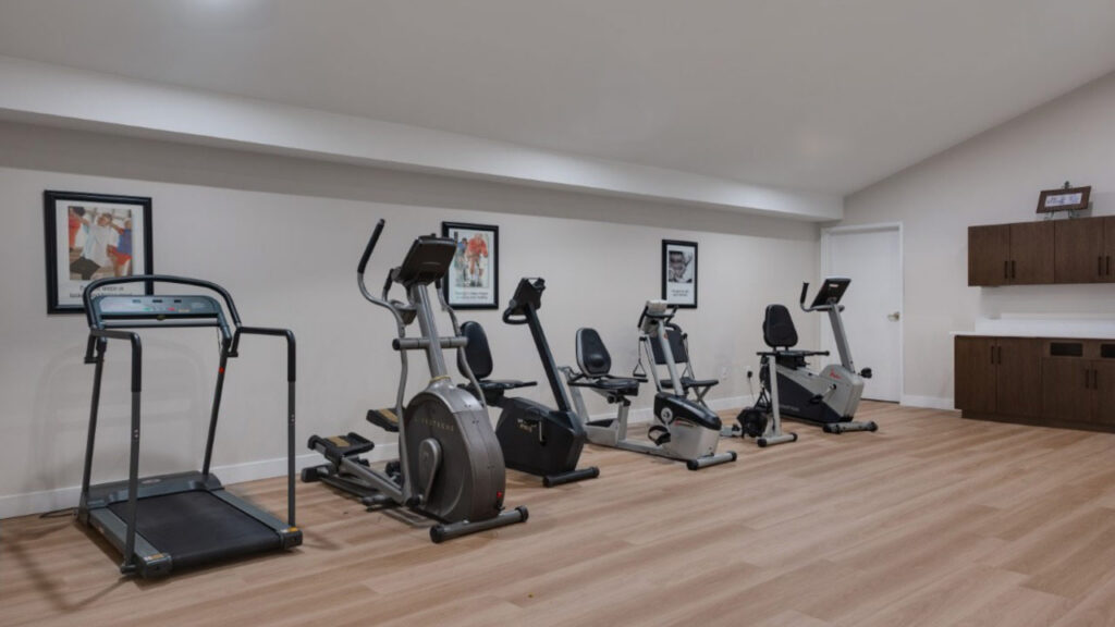 A home gym room with a treadmill, three stationary bikes, wooden flooring, white walls with sports-themed pictures, and brown cabinets on the right side.