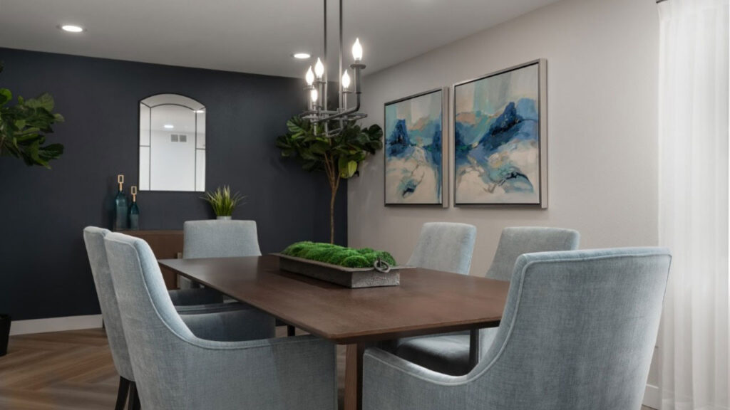 Modern dining room with a wooden table, six light blue upholstered chairs, two abstract paintings, a black chandelier, and potted plants. A window with white curtains lets in natural light.