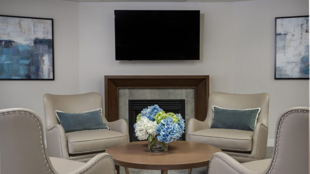 Two beige armchairs with blue pillows face a round table with a blue and white flower arrangement. A flat-screen TV and two abstract paintings hang on the light gray walls above a fireplace.