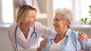 A healthcare provider with a stethoscope smiles and talks to an elderly woman, gently touching her shoulder, in a bright, comfortable room, demonstrating compassionate health management in senior living.
