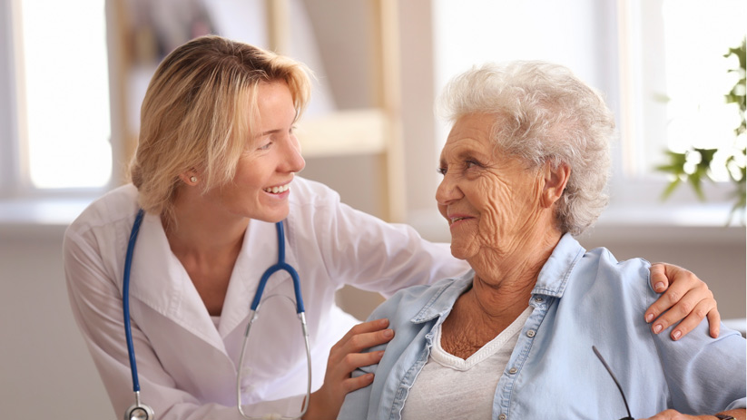 A healthcare provider with a stethoscope smiles and talks to an elderly woman, gently touching her shoulder, in a bright, comfortable room, demonstrating compassionate health management in senior living.