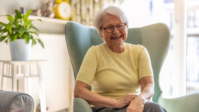 An older woman with short gray hair and glasses sits in a pale green armchair, smiling at the camera while practicing effective health management in senior living.