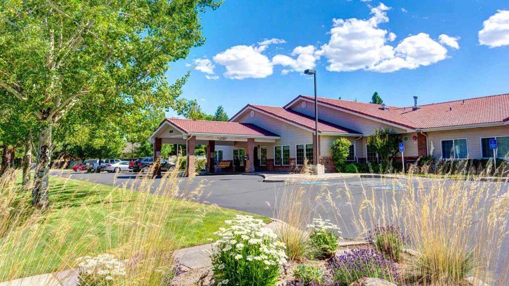 A single-story Memory Care building with a red roof sits beside a parking lot, surrounded by green trees, tall grasses, and blooming white flowers under a blue sky with scattered clouds.